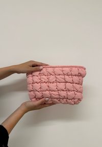 Pochette en tissu matelassé rose, de forme rectangulaire, avec un bord supérieur plissé et une texture douce et rembourrée.