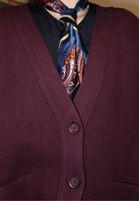 Bordový kardigán s výstrihom do V, textúrovanou látkou, dvoma gombíkmi vpredu a vreckom; kombinovaný s vzorovaným hodvábnym šálom previazaným okolo krku.