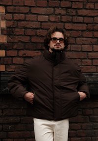 Brown puffer jacket with a high collar, zippered pockets, and a smooth texture, paired with light-coloured trousers against a brick wall.
