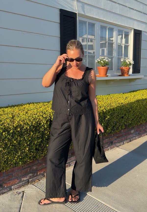 Žena v čiernom nátelníku a nohaviciach so slnečnými okuliarmi, drží čiernu tašku, stojí na chodníku pred bielym domom a živým plotom.