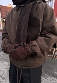 Ruda pūkinė striukė su austa rankogaliais, tamsiai rudos vilnonės skaros su kutais kraštais ir rudos pirštinės su tekstūruotais elementais.