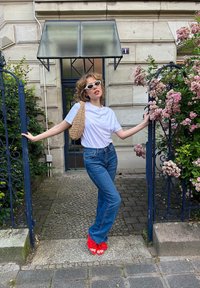 T-shirt blanc, jeans taille haute bleus, sandales à fleurs rouges et sac en osier, se tenant près d'une porte en métal avec des fleurs roses en arrière-plan.