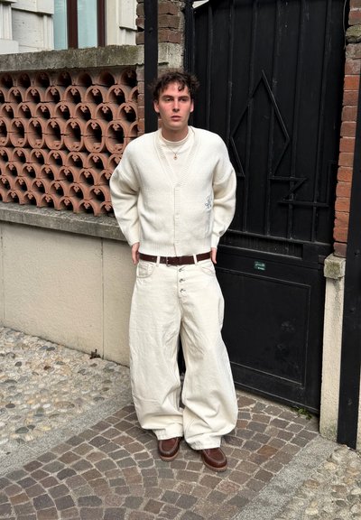Joven de pie sobre pavimento adoquinado con pantalones blancos anchos, cárdigan blanco, cuello alto, cinturón marrón y zapatos marrones junto a una puerta negra.