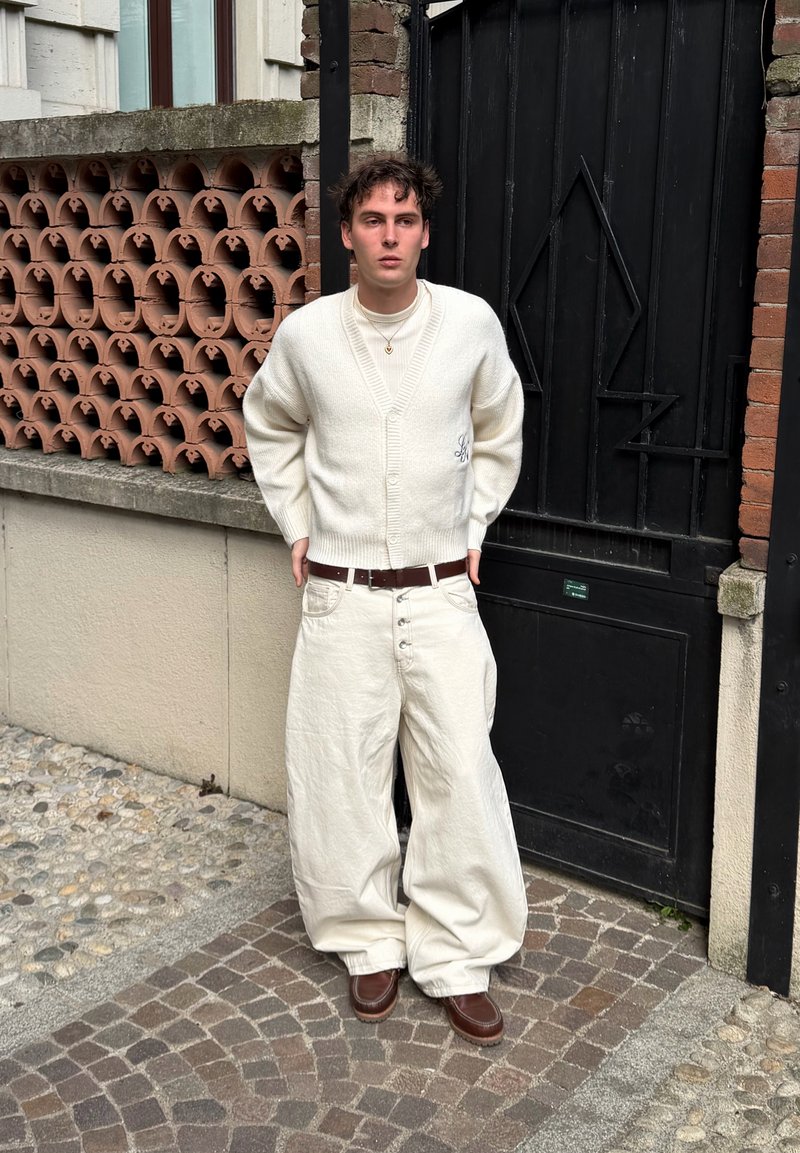 Ένας νεαρός άντρας στέκεται σε πλακόστρωτο πεζοδρόμιο, φοράει λευκό φαρδύ παντελόνι, λευκή ζακέτα, ζιβάγκο, καφέ ζώνη και καφέ παπούτσια δίπλα σε μαύρη πόρτα.