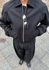 Crna bomber jakna sa zatvaračem, raglan rukavima i elastičnim manžetama. Uparena s labavim crnim hlačama i sjajnim gležnjačama.