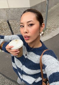 Flauschige, gestreifte Strickjacke in Marineblau und Grau. Weicher Stoff, mit Knöpfen vorne, getragen mit einem braunen Pappbecher. Ein brauner Handtaschenriemen ist sichtbar.