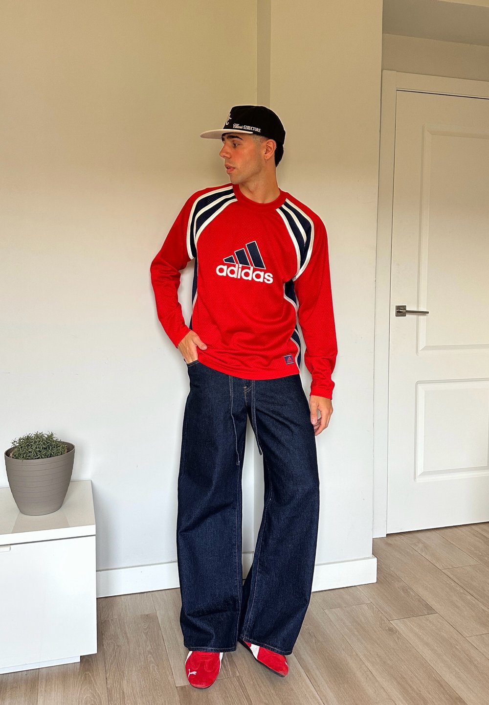 Rød langermet Adidas-skjorte med marineblå aksenter, kombinert med vide mørkeblå jeans og røde sko. Enkel bakgrunn med en potteplante.