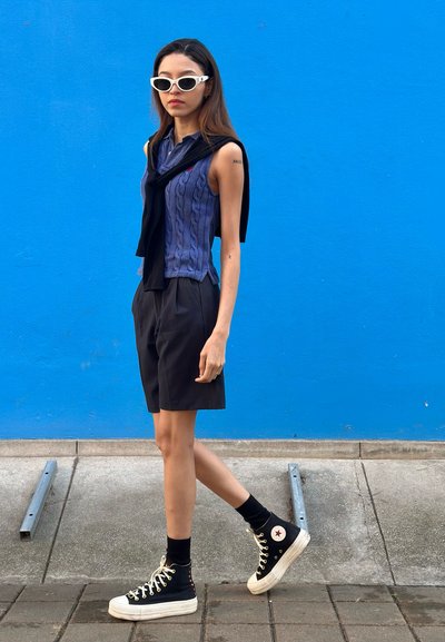 Modelo que lleva un top sin mangas de punto en color púrpura, pantalones cortos negros y zapatillas altas negras, con un suéter negro caído sobre los hombros.