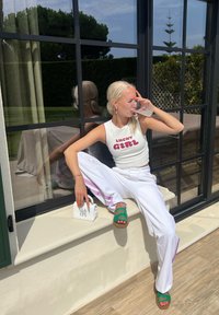 Ξανθιά γυναίκα με λευκή μπλούζα "Lucky Girl" και λευκά παντελόνια κάθεται κοντά σε παράθυρο, πίνει από ποτήρι, κρατώντας μια μικρή λευκή τσάντα.