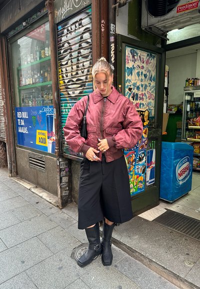 Chaqueta bomber burdeos, con corte oversized, puños elásticos, combinada con culottes negros y botas de goma negras gruesas, de pie delante de una tienda.
