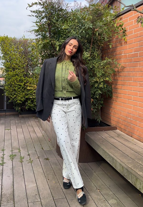 O femeie stă pe o terasă din lemn, purtând un cardigan verde, un sacou închis la culoare, pantaloni albi cu buline și balerini negri, lângă un perete de cărămidă și plante.