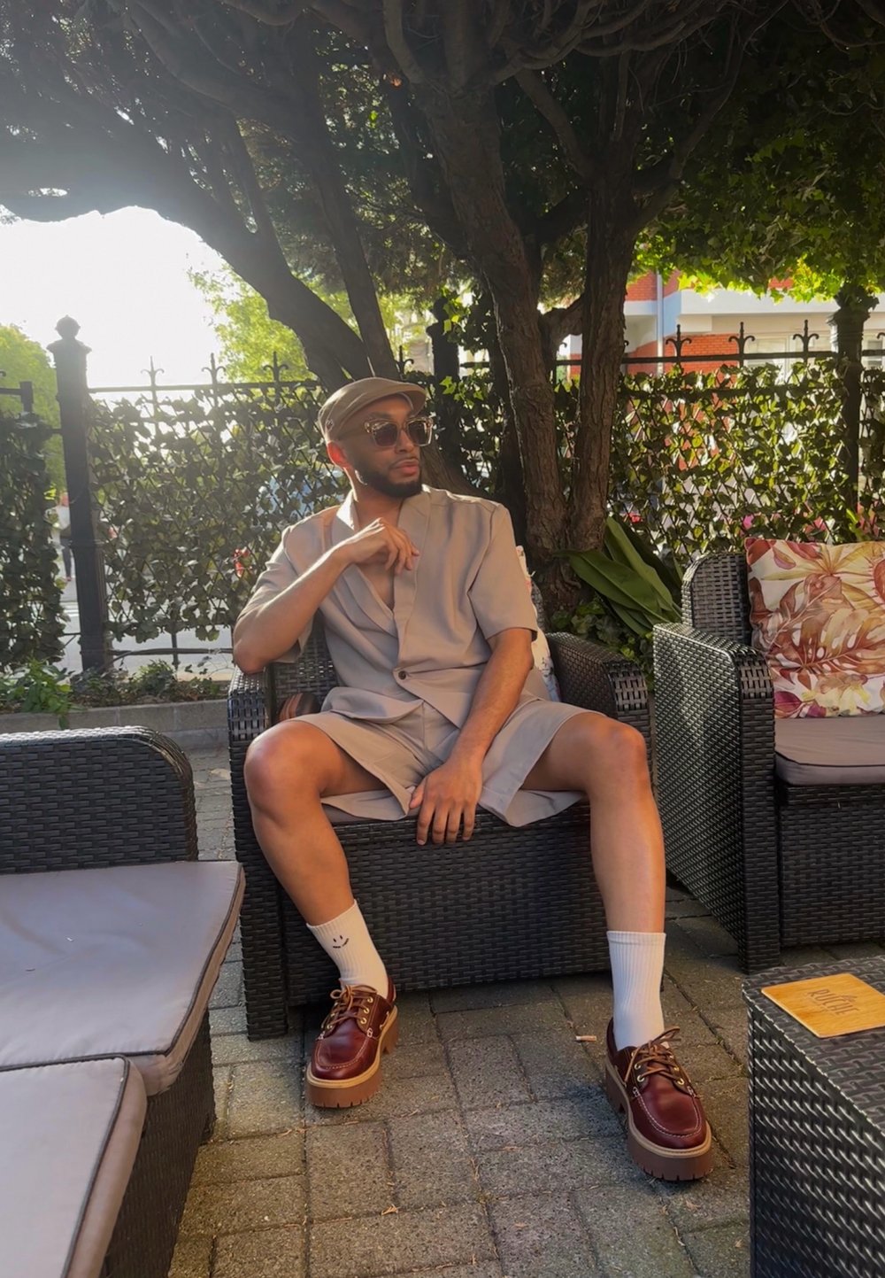 Ανοιχτό γκρι σακάκι κοντό μανίκι με χαλαρή εφαρμογή, συνδυασμένο με άσπρες κάλτσες και καφέ παπούτσια με πλατφόρμα. Καθισμένος σε καρέκλα από ρατάν σε εξωτερικό χώρο.