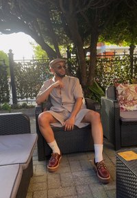 Ανοιχτό γκρι σακάκι κοντό μανίκι με χαλαρή εφαρμογή, συνδυασμένο με άσπρες κάλτσες και καφέ παπούτσια με πλατφόρμα. Καθισμένος σε καρέκλα από ρατάν σε εξωτερικό χώρο.