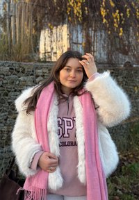 Giacca in pelliccia sintetica bianca sopra una felpa rosa con la scritta "SPY" stampata e una sciarpa rosa chiaro con frange. Ambientazione all'aperto con fogliame sullo sfondo.