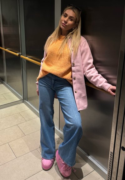 Joven con cabello largo y rubio, vestida con un suéter naranja, abrigo rosa, jeans azules y zapatos rosas, apoyada contra una pared de metal en un interior.