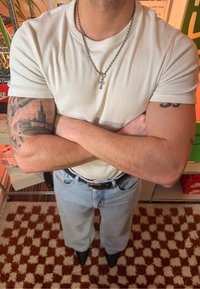 T-shirt blanc côtelé, jean large bleu clair, ceinture noire et chaussures pointues noires. Tatouages visibles sur les bras. En arrière-plan, des étagères.