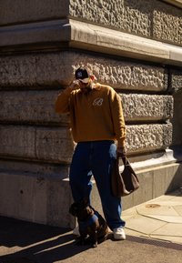 Pull marron en tricot avec un col et un logo "B", jean bleu, et un sac sombre. Un chien noir avec un harnais est assis à proximité.