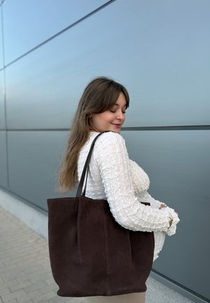 Junge Frau in einem strukturierten weißen Pullover, die eine große dunkelbraune Tragetasche hält, steht draußen vor einer grauen Wand, die Augen geschlossen, lächelt.