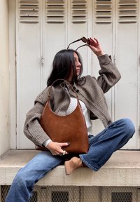 Geantă hobo din piele maro, cu textură netedă, formă semi-circulară. Modelul poartă o jachetă houndstooth, blugi albaștri și botine bej.
