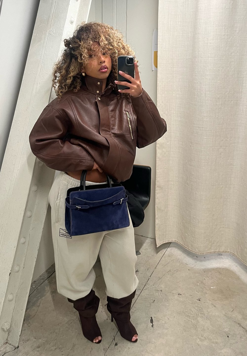 Femme aux cheveux bouclés portant une veste en cuir marron oversize, un pantalon blanc, des bottines à bouts ouverts marron foncé, tenant un sac à main bleu marine, prenant un selfie dans le miroir.