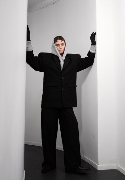 Traje negro de gran tamaño con hombros acolchados, sudadera gris por debajo y guantes de cuero negro; se apoya contra una pared blanca en un espacio estrecho.