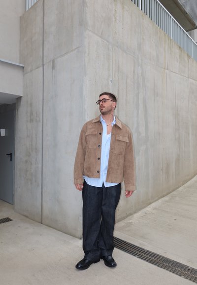 Hombre con chaqueta de ante marrón, camisa azul clara, pantalones de rayas y zapatos negros, de pie junto a una gran pared de hormigón al aire libre.