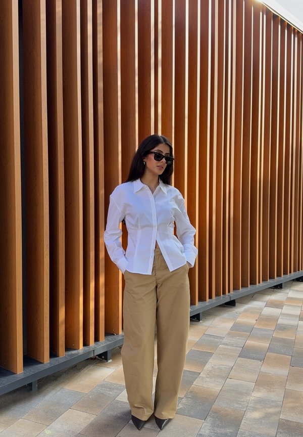 Una mujer con gafas de sol, camisa blanca y pantalones beige está de pie con las manos en los bolsillos junto a una pared de listones de madera verticales.