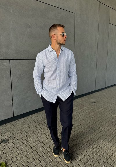 Camisa de botones a rayas azul y blanca, pantalones navy y alpargatas negras. De pie contra una pared gris, con las manos en los bolsillos. Diseño casual.