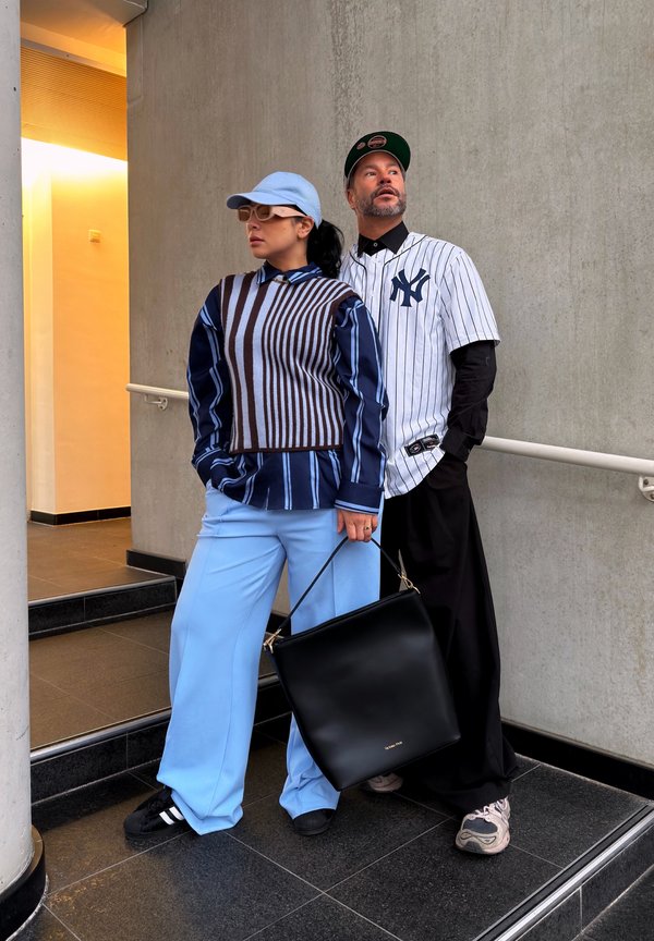 Two people stand indoors against a concrete wall; one wears light blue trousers and a striped vest, the other a white pinstriped baseball jersey.