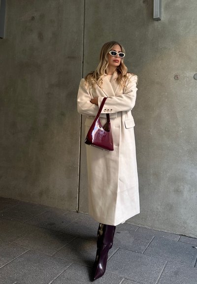 Abrigo de mezcla de lana beige con un diseño de doble botonadura, combinado con un bolso burdeos brillante y botas de tacón puntiagudas en un tono similar.