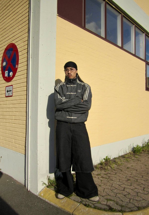 Person trägt eine graue Adidas-Jacke und weite schwarze Hosen und steht mit verschränkten Armen vor einer gelben Backsteinmauer in der Nähe eines Parkverbotszeichens.