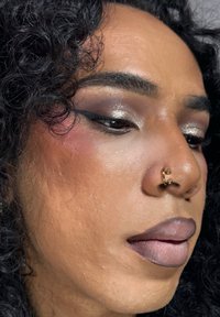Nærbilde av et ansikt med krøllete svart hår, med en gull septumring, markert eyeliner og matt, dempet leppestift. Hudtekstur synlig.