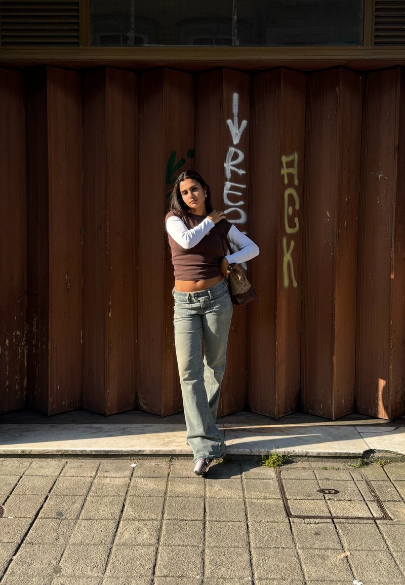 Jeune femme en haut superposé et jean se tient sur le trottoir de la ville devant un mur ondulé marron avec des graffitis au soleil.