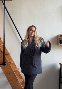 Sacou negru cu două rânduri de nasturi, dotat cu două buzunare frontale, purtat alături de pantaloni negri asortați. Scări de lemn și decor minimalist în fundal.