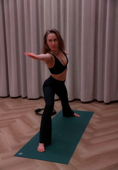 Mujer con ropa deportiva negra realizando una postura guerrera de yoga sobre una esterilla verde en una habitación con suelo de madera y cortinas beige.
