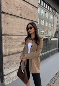 Ανοιχτό καφέ σουέτ σακάκι με χαλαρή εφαρμογή, μαύρη βερμούδα, λευκό μπλουζάκι και σκούρο καφέ τσάντα; φορώντας μαύρα γυαλιά ηλίου και ένα κολιέ.