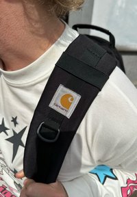 Correia de mochila preta com um patch com o logo da Carhartt em amarelo e branco, fabricada com material resistente e detalhe de fivela ajustável.