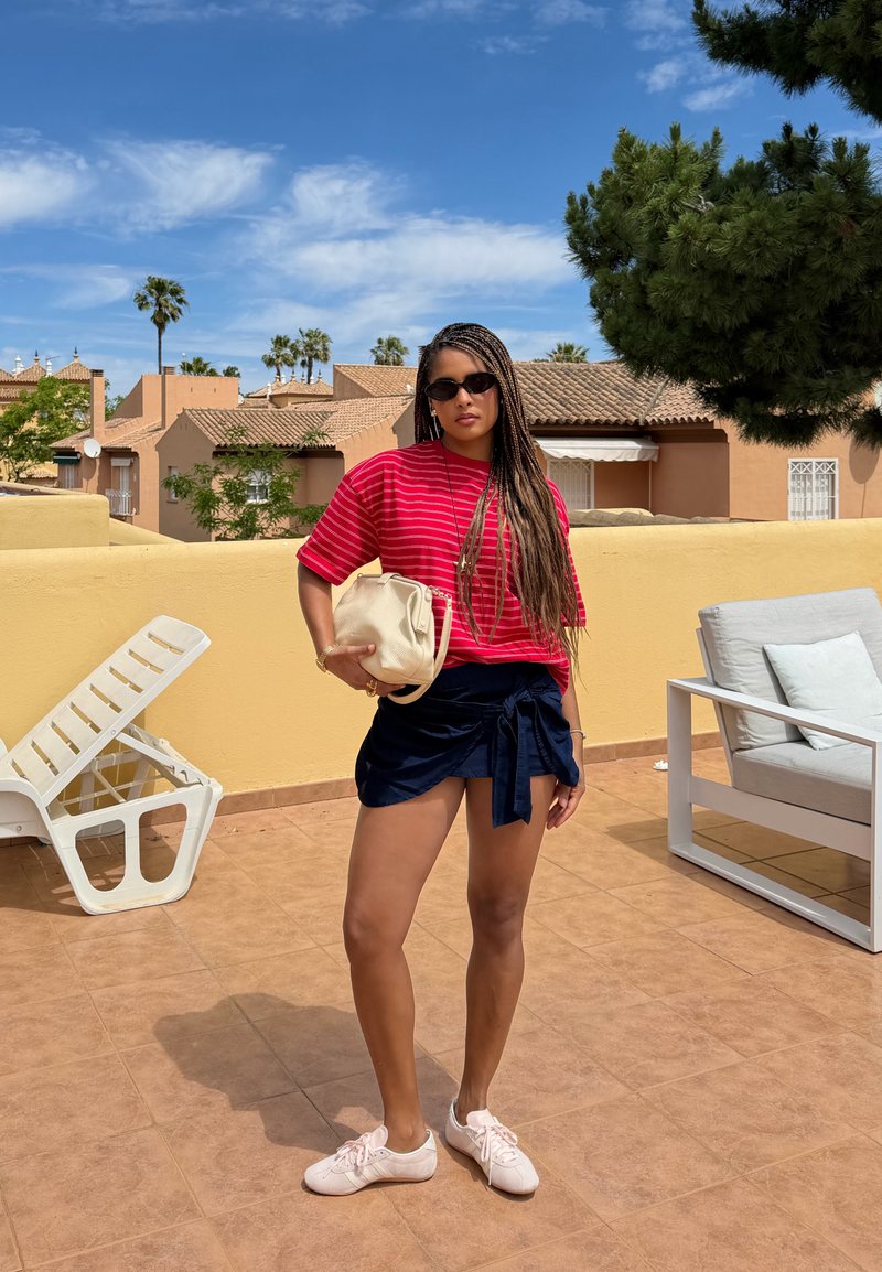 Femme aux longs cheveux tressés portant des lunettes de soleil, une chemise rouge rayée, une jupe portefeuille bleu marine et des baskets blanches, debout sur une terrasse avec mobilier de jardin.