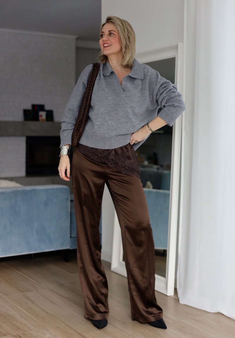Donna in piedi all'interno che indossa un maglione grigio, pantaloni in satin marrone con dettagli in pizzo, scarpe nere con punta e una borsa intrecciata marrone portata a tracolla.