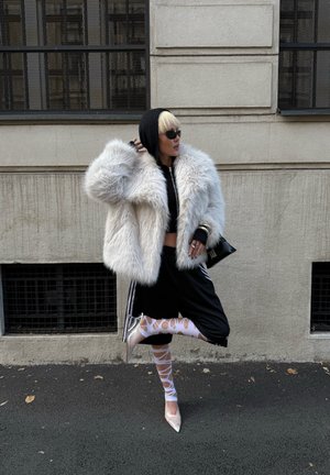 Abrigo de peluche blanco, sudadera negra con capucha, top corto, pantalones deportivos negros con rayas blancas; medias blancas transparentes, tacones transparentes, gafas de sol.