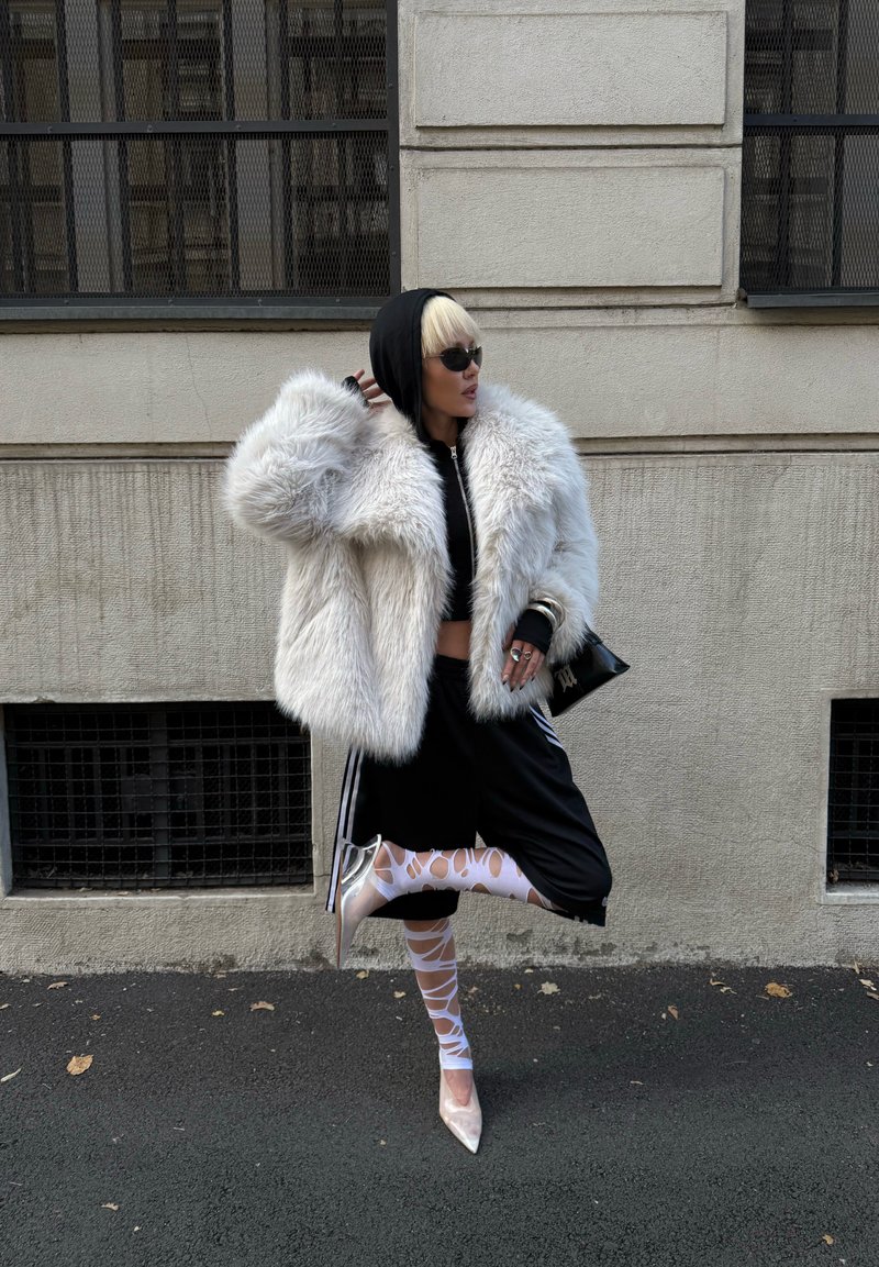 White faux fur coat, black hoodie, cropped top, black athletic pants with white stripes; sheer white tights, transparent heels, sunglasses.