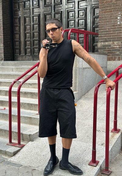 Hombre con camiseta sin mangas negra y pantalones cortos grabando con una videocámara, apoyado en pasamanos rojos cerca de escalones de piedra y puertas de madera oscura.
