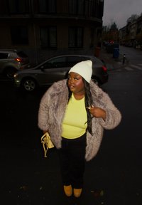 Manteau en fausse fourrure gris clair, chemise jaune fluo, pantalon noir et chaussures à plateforme jaunes. Un bonnet blanc est porté, avec un petit sac à main jaune.