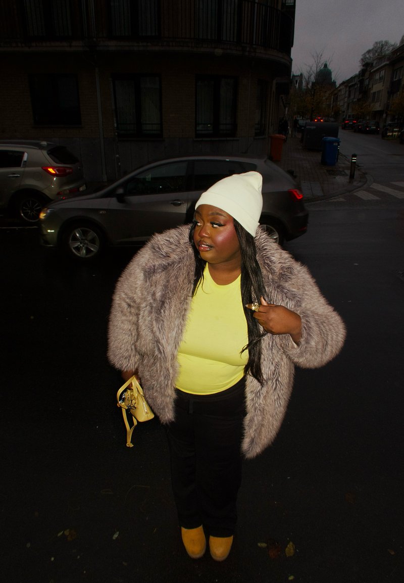 Manteau en fausse fourrure gris clair, chemise jaune fluo, pantalon noir et chaussures à plateforme jaunes. Un bonnet blanc est porté, avec un petit sac à main jaune.