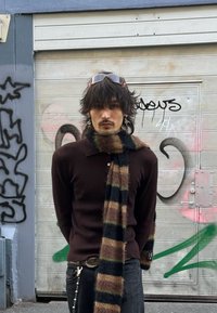 Pull en maille marron avec une texture côtelée et un col, associé à une écharpe rayée en marron, vert et noir. Pantalon noir avec un détail en chaîne.