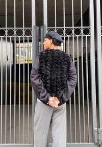 Black faux fur vest layered over a long-sleeve black top; grey trousers; a person standing in front of a metal gate with vertical bars.