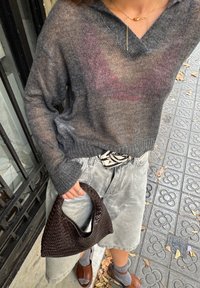 Šedý, průsvitný pletený svetr s výstřihem do V, spárovaný se světle šedými, texturovanými šortkami. Drží tmavou tkanou kabelku s zakřiveným uchem.