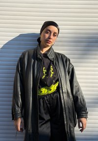 Femme portant une veste en cuir noire, un foulard noir sur la tête et un t-shirt graphique Adidas, debout contre un mur blanc texturé au soleil.