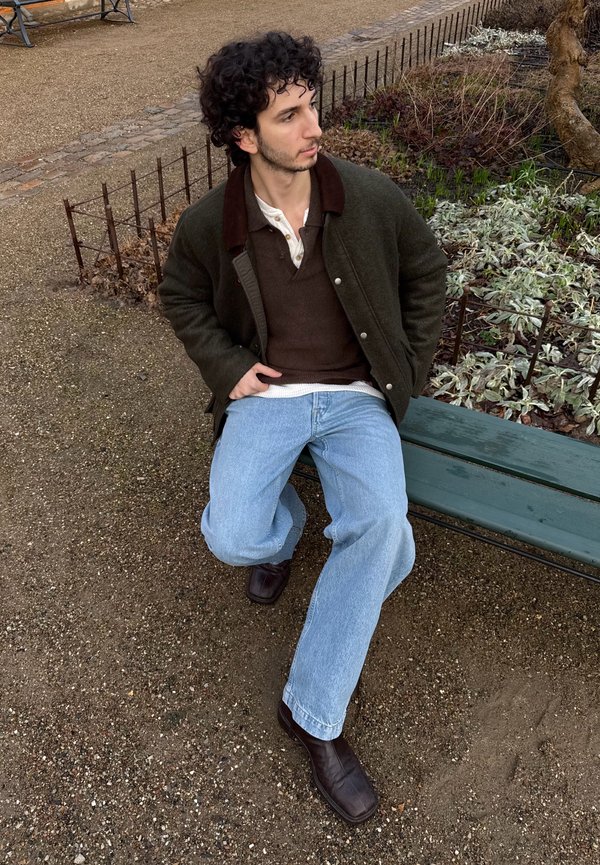 Homme aux cheveux bouclés portant une veste marron, un pull foncé et un jean clair, assis sur un banc vert dans un parc, près de plantes et d'un chemin de gravier.