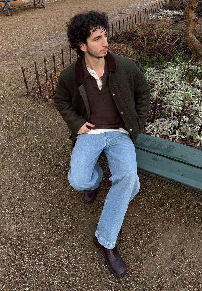 Homme aux cheveux bouclés portant une veste marron, un pull foncé et un jean clair, assis sur un banc vert dans un parc, près de plantes et d'un chemin de gravier.